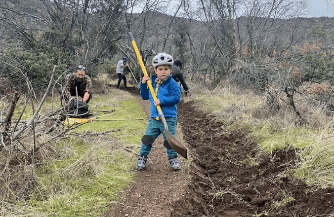 Un niño con casco levanta una pala junto a voluntarios trabajando en un sendero de tierra